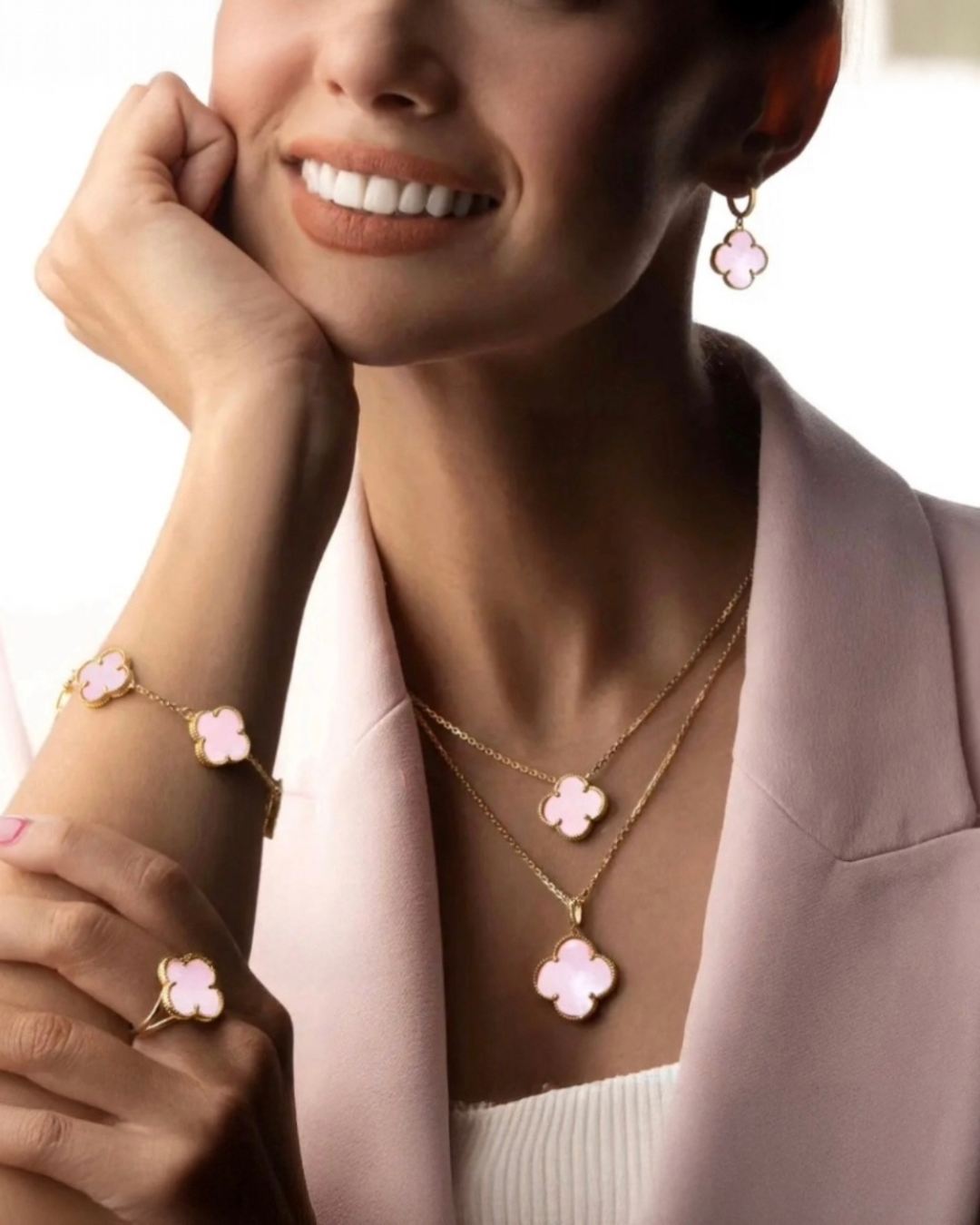 Elegantes Schmuck-Set aus 18k Gelbgold mit rosafarbenen Kleeblatt-Anhängern – Halsketten im Layering-Look, Armband, Ring und Ohrringe mit zartem Rosa-Inlay, stilvoll getragen zu einem hellrosa Blazer, luxuriöse feminine Ausstrahlung.