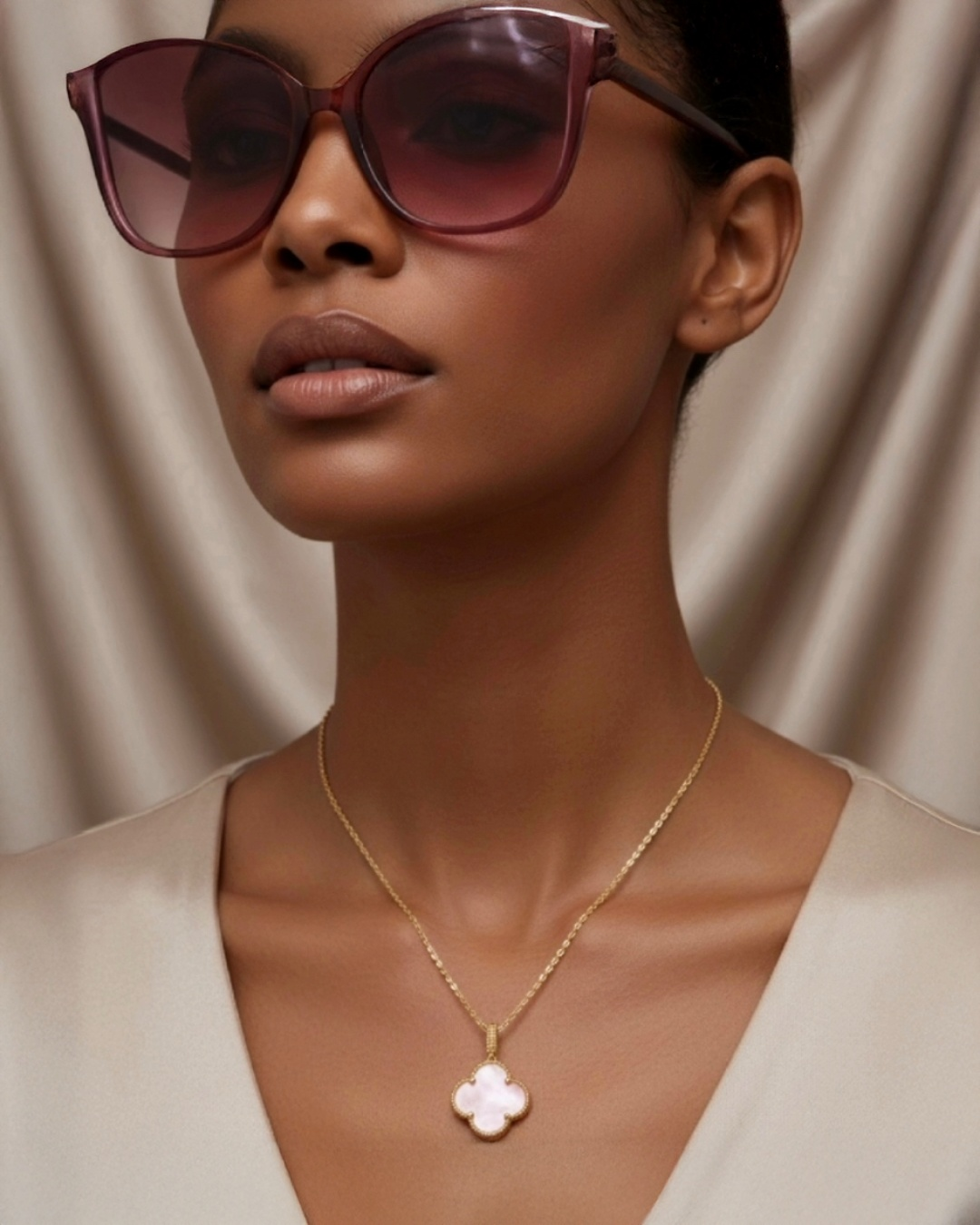 Elegantes Nahporträt einer Frau mit oversized, transparent-rosafarbenen Sonnenbrillen. Sie trägt eine feine 18k-Gelbgold-Halskette mit einem weißen, kleeblattförmigen Anhänger in Perlmutt-Optik. Dazu kombiniert sie ein beiges V-Ausschnitt-Oberteil vor einem weich fallenden, neutralen Stoffhintergrund, der die warmen Haut- und Goldtöne betont und eine luxuriöse, minimalistische Atmosphäre schafft.