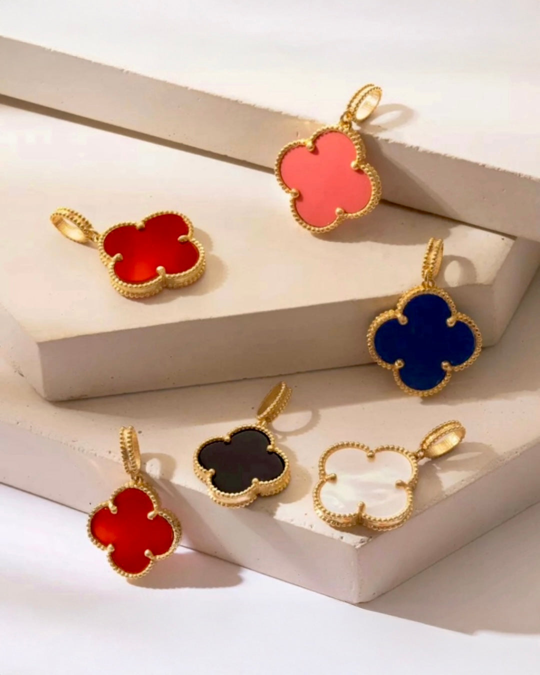 Mehrere kleeblattförmige Anhänger aus 18 Karat Gelbgold mit farbigen Einlagen in Rot, Rosa, Blau, Schwarz und Weiß, arrangiert auf gestuften, hellen Podesten.