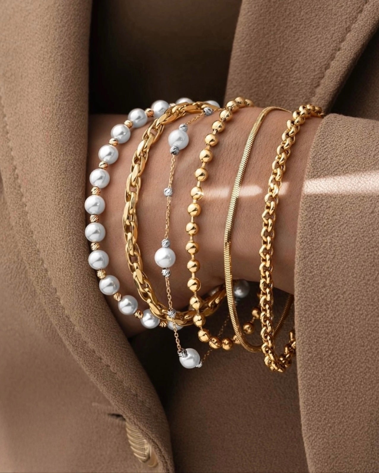 Mehrere Armbänder aus 18 Karat Gelbgold, kombiniert mit weißen Perlenarmbändern – darunter Ketten-, Glieder- und Kugeldesigns – elegant am Handgelenk getragen und stilvoll mit einem beigefarbenen Blazer kombiniert.