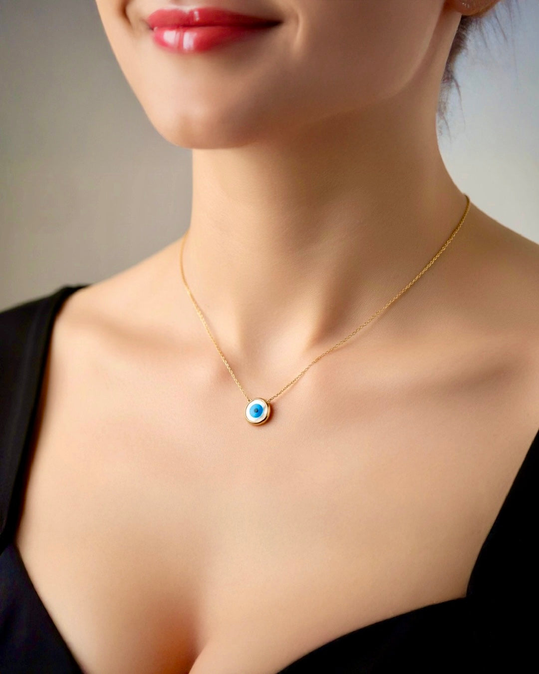 Goldene, filigrane Halskette mit rundem Anhänger im Nazar-Design (blau-weisses Auge) getragen am Hals einer Frau mit schwarzem Oberteil; weiches Licht, neutraler Hintergrund, Fokus auf dem Schmuckstück.