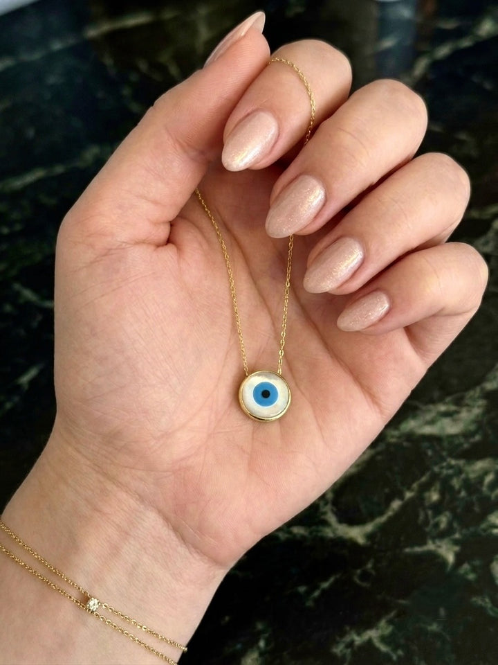 Nahaufnahme einer Hand mit dezent schimmernd lackierten, mandelförmigen Fingernägeln, die eine filigrane Halskette aus Gelbgold hält. Im Mittelpunkt hängt ein rundes Nazar-Auge-Amulett mit goldfarbenem Rand sowie weißen, hellblauen, dunkelblauen und schwarzen Kreisen. Im Hintergrund ist eine dunkle Marmoroberfläche zu sehen. Am Handgelenk trägt die Person zusätzlich zwei feine Goldarmbänder, eines davon mit einem kleinen funkelnden Stein.