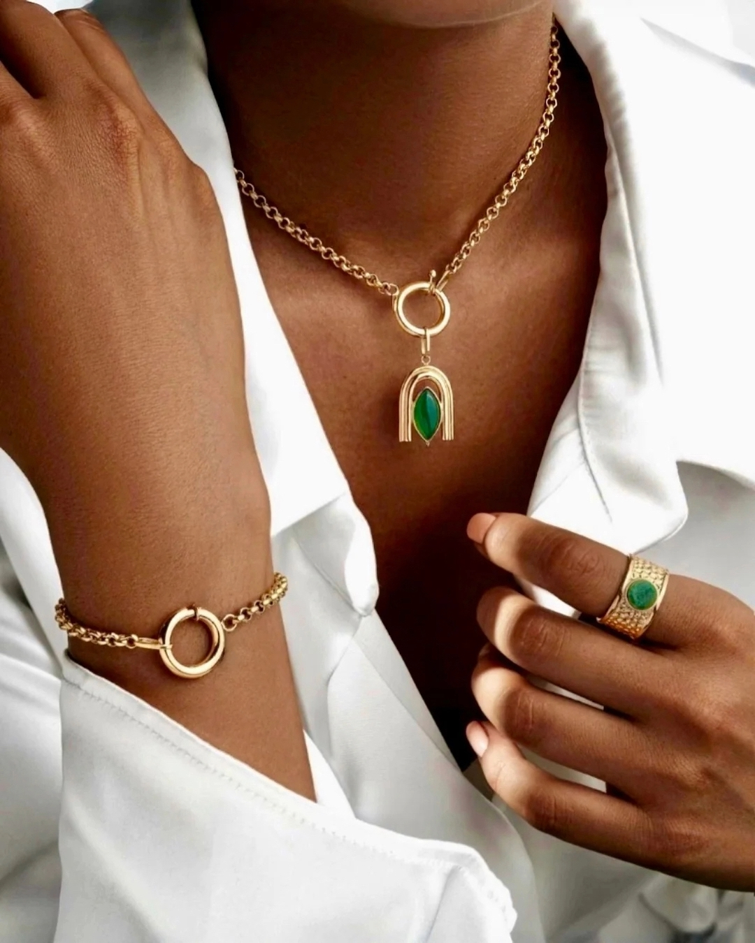Elegante Frau trägt eine feine 18k-Gelbgoldkette mit Anhänger und grünem Stein, dazu passendes Goldarmband und Goldring mit grünem Edelstein, stilvoll kombiniert zu weißer Bluse.