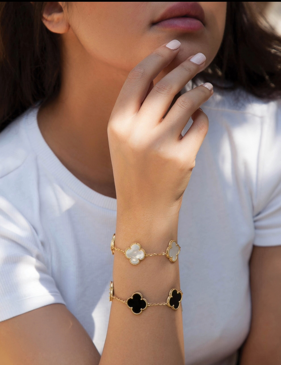 Handgelenk mit zwei filigranen Armbändern aus 18k Gelbgold, eines mit schwarzem Onyx und eines mit hellem Perlmutt in kleeblattförmigem Design, elegant getragen bei Tageslicht.
