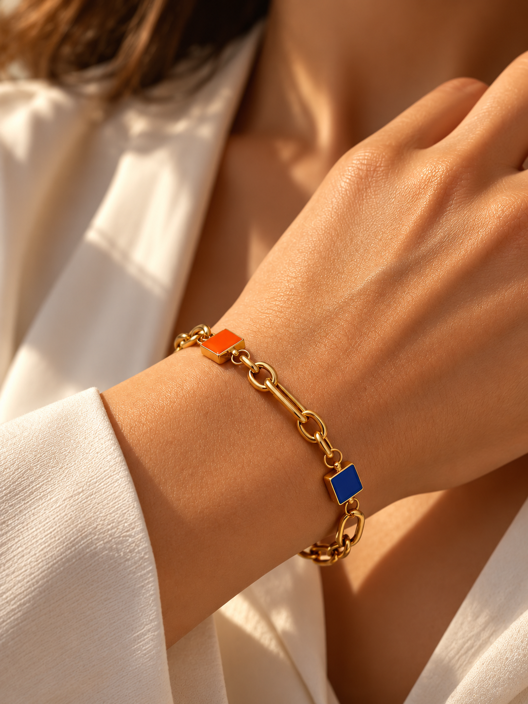 Elegantes Goldarmband mit ovalen Kettengliedern und zwei quadratischen Schmuckelementen in Orange und Blau am Handgelenk einer Frau, stilvoll kombiniert mit einer weissen Bluse im warmen Sonnenlicht.