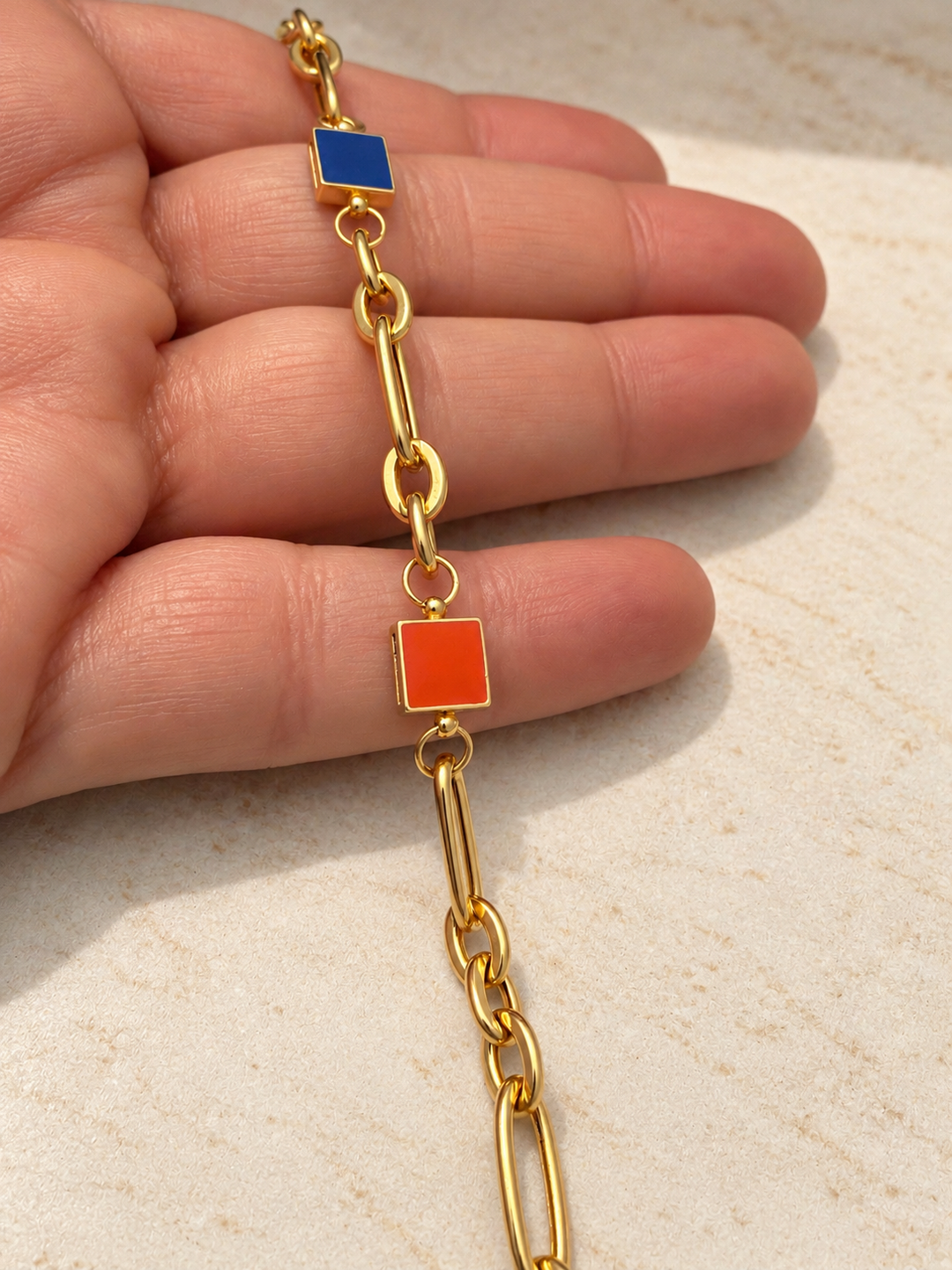 Goldarmband aus 18k Gelbgold mit länglichen Kettengliedern und zwei quadratischen Elementen in Orange und Blau auf heller Unterlage.
