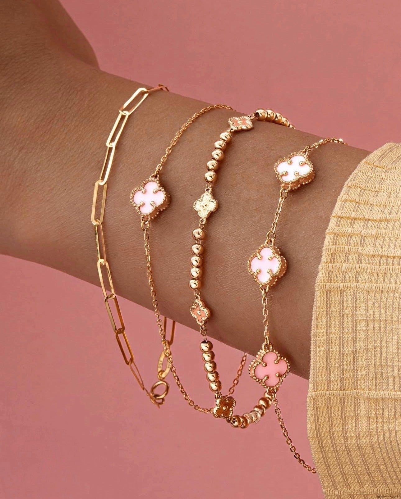 Nahaufnahme mehrerer übereinander getragener Armbänder aus 18k Gelbgold. Das mittlere Armband („Reshteh“ – Verbindung) kombiniert polierte Goldperlen mit zarten Kettengliedern und kleinen, rosafarben emaillierten Kleeblatt-Elementen. Daneben ein Armband mit länglichen Gliedern sowie ein weiteres mit ausschließlich Kleeblatt-Motiven. Hintergrund in sanftem Rosaton, getragen zu einem beigefarbenen Oberteil.