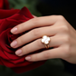 Eleganter 18k Gelbgold-Ring mit Kleeblatt-Motiv aus weißem Perlmutt, getragen an einer Hand vor einer roten Rose; luxuriöser, zeitloser Schmuck mit filigraner Goldfassung und natürlicher Steinstruktur.