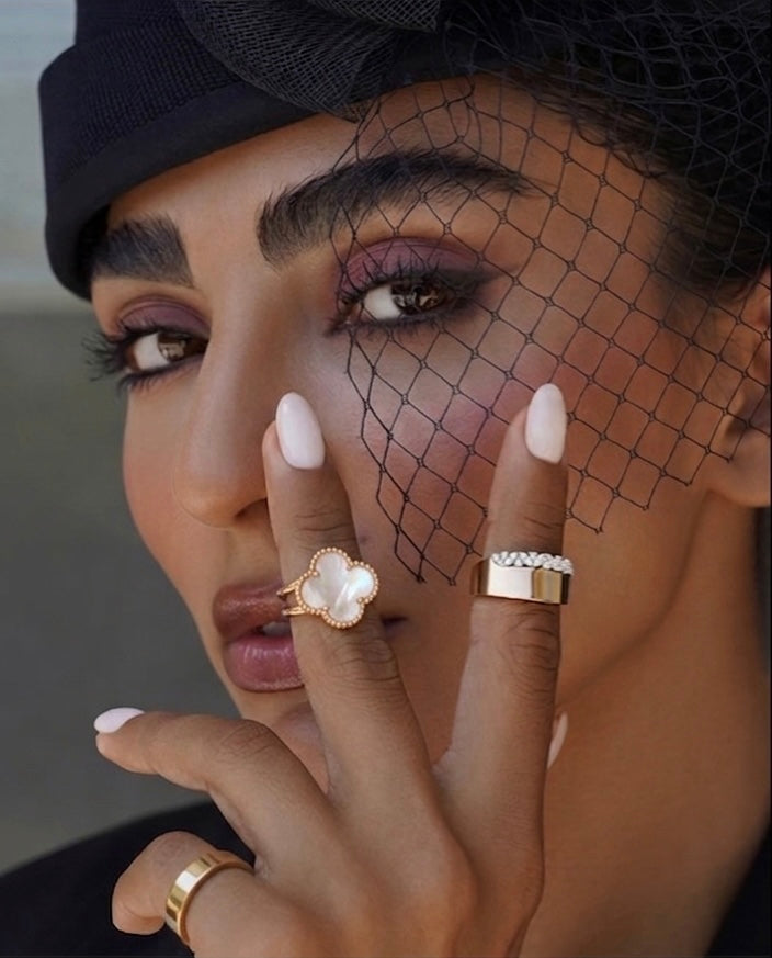 Elegantes Beauty-Porträt einer Frau mit schwarzem Fascinator und feinem Gesichtsschleier. Sie trägt mehrere moderne Goldringe, darunter einen auffälligen Ring mit vierblättrigem, perlmuttfarbenem Motiv sowie schlichte, hochglanzpolierte Goldringe. Das Make-up mit violettem Lidschatten und die hellen Nägel unterstreichen den luxuriösen, stilvollen Look. Die Hand ist kunstvoll vor dem Gesicht positioniert und lenkt den Fokus auf den Schmuck und die ausdrucksstarken Augen.
