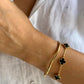 Ansicht eines Handgelenks mit zwei Armbändern: einem schlichten, geschwungenen Armreif aus Gelbgold und einem filigranen Goldarmband mit schwarzen, vierblättrigen Onyx-Elementen, getragen zu einer weißen Bluse.