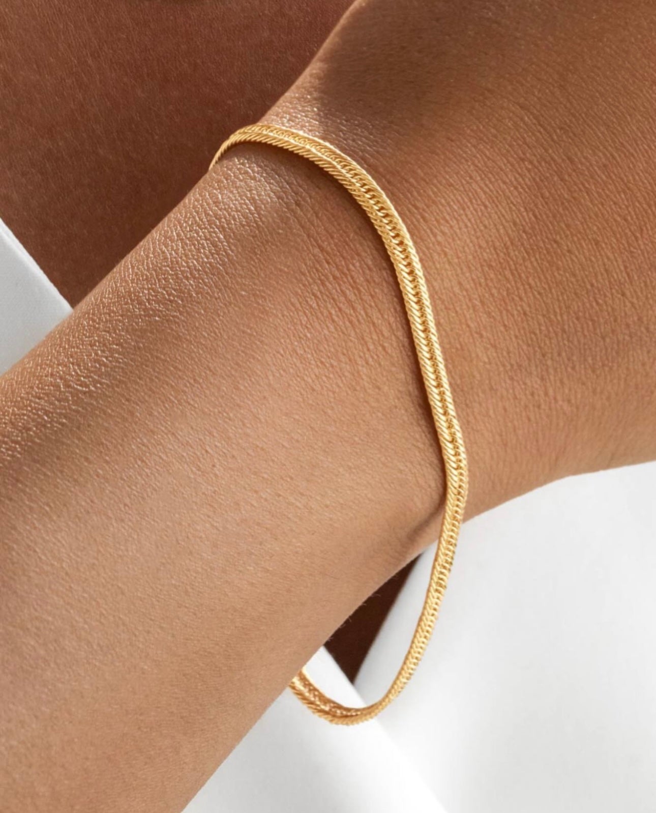 Feines goldenes Armband mit flach anliegender Gliederstruktur, locker um das Handgelenk einer Person mit gebräunter Haut gelegt, vor weißem Hintergrund.