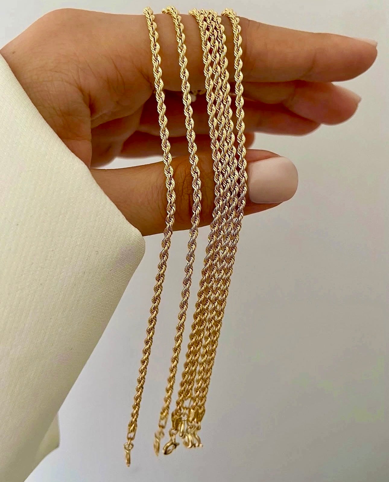 Nahaufnahme mehrerer Armbänder aus 18k Gelbgold in gedrehter Kordeloptik, die locker über die Finger einer Hand gelegt sind. Die polierten, spiralförmig verflochtenen Glieder reflektieren das Licht und erzeugen einen intensiven Goldglanz. Hintergrund neutral und hell, Hand in weißem Stoffärmel.