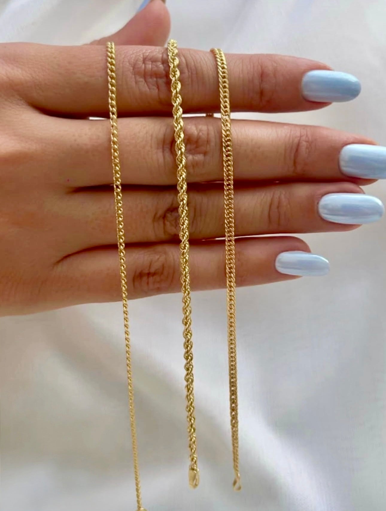 Drei feine Armbänder aus 18k Gelbgold auf einer Hand mit hellblau lackierten Nägeln – links schlicht, in der Mitte gedreht als goldene Kordel (Tanab-e Tala), rechts flachgliedrig.