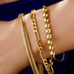 Drei stilvolle Goldarmbänder an einem Handgelenk: ein markantes Spiralarmband im Coil-Design, ein modernes Gliederarmband sowie ein offener Armreif mit hochglanzpolierten Goldkugeln. Die Schmuckstücke wirken elegant und luxuriös vor dunklem Hintergrund.