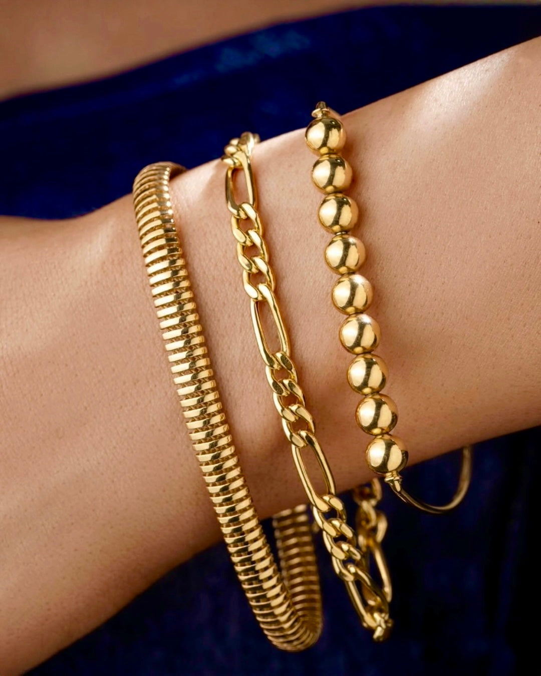 Drei stilvolle Goldarmbänder an einem Handgelenk: ein markantes Spiralarmband im Coil-Design, ein modernes Gliederarmband sowie ein offener Armreif mit hochglanzpolierten Goldkugeln. Die Schmuckstücke wirken elegant und luxuriös vor dunklem Hintergrund.