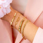 Elegante Goldarmbänder in verschiedenen Designs an einem Handgelenk vor zartrosa Hintergrund. Zu sehen sind ein geometrisches Armreif-Design mit quadratischen Elementen, ein feines Kettenarmband mit ovalem Detail, ein modernes Spiral-Armreif sowie ein Gliederarmband in glänzendem Goldton.