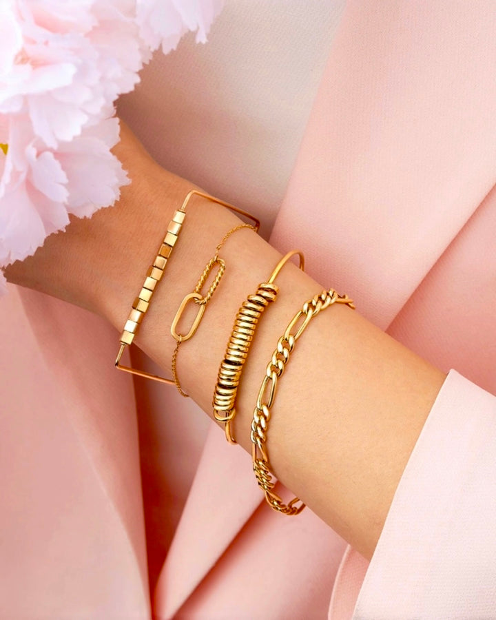 Elegante Goldarmbänder in verschiedenen Designs an einem Handgelenk vor zartrosa Hintergrund. Zu sehen sind ein geometrisches Armreif-Design mit quadratischen Elementen, ein feines Kettenarmband mit ovalem Detail, ein modernes Spiral-Armreif sowie ein Gliederarmband in glänzendem Goldton.