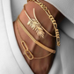 Eleganter Armschmuck aus Gold am Handgelenk, bestehend aus einem polierten Figaro-Armband, einem offenen Armreif mit filigranem Blattdesign, einem geometrischen Goldarmband sowie einem feinen Kettenarmband mit ovalen Gliedern. Stilvoll kombiniert mit einem weißen Blazer für einen luxuriösen und modernen Look.
