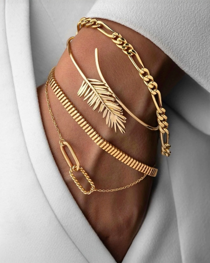 Eleganter Armschmuck aus Gold am Handgelenk, bestehend aus einem polierten Figaro-Armband, einem offenen Armreif mit filigranem Blattdesign, einem geometrischen Goldarmband sowie einem feinen Kettenarmband mit ovalen Gliedern. Stilvoll kombiniert mit einem weißen Blazer für einen luxuriösen und modernen Look.