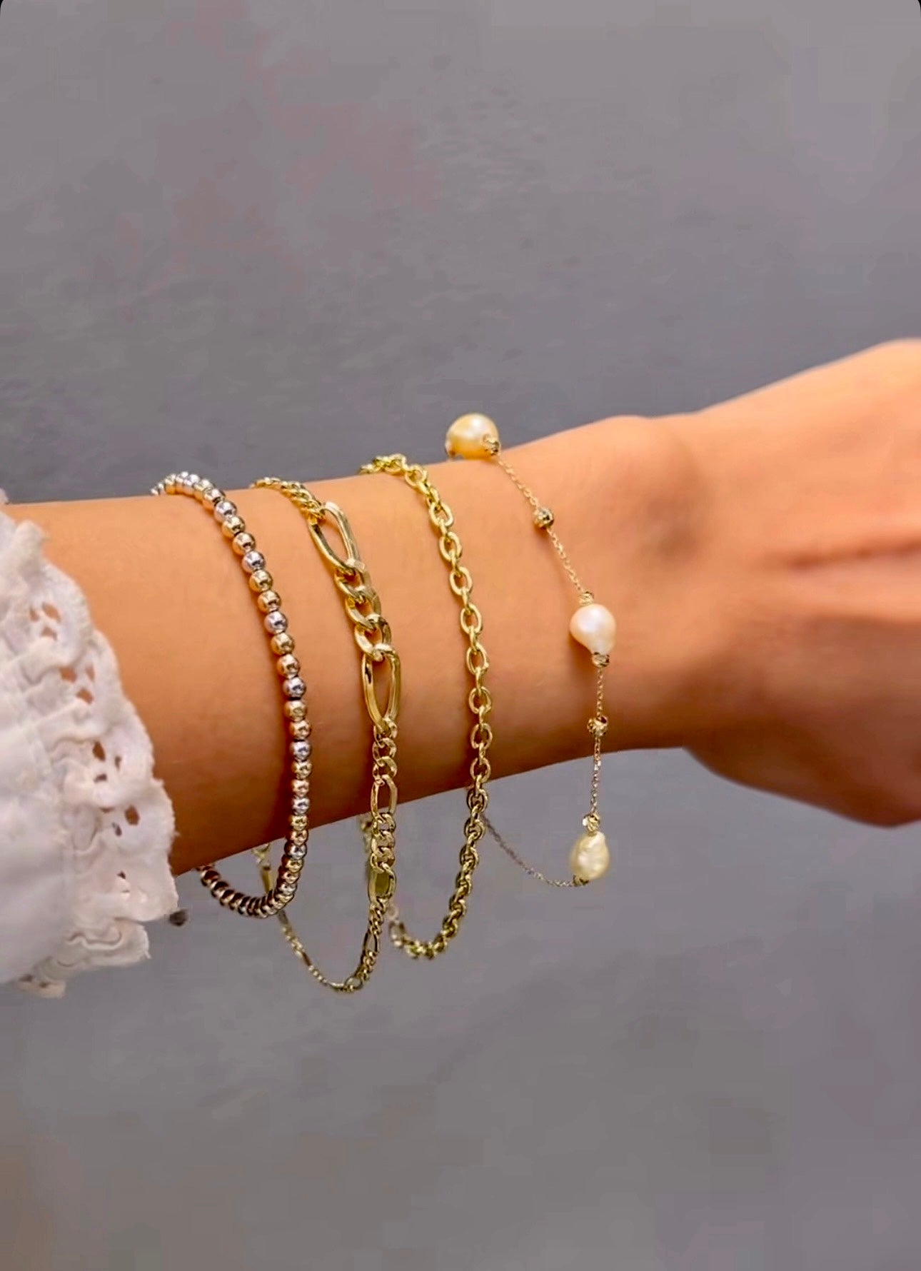 Nahaufnahme eines 18k Gelbgold-Armbands im Figaro-Conic-Design, getragen am Handgelenk zusammen mit weiteren Armbändern aus Gold, Perlen und Silber, vor neutralem Hintergrund.