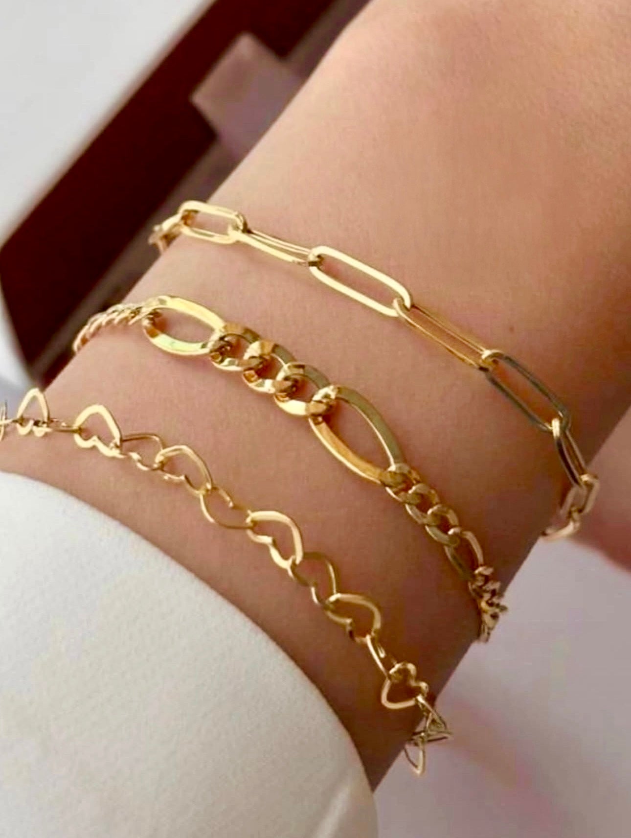 Drei goldene Armbänder am Handgelenk einer Person mit heller Haut, bestehend aus einem Armband mit länglichen Kettengliedern, einem Figaro-Gliederarmband mit abwechselnd großen und kleinen Gliedern sowie einem filigranen Armband mit herzförmigen Gliedern.