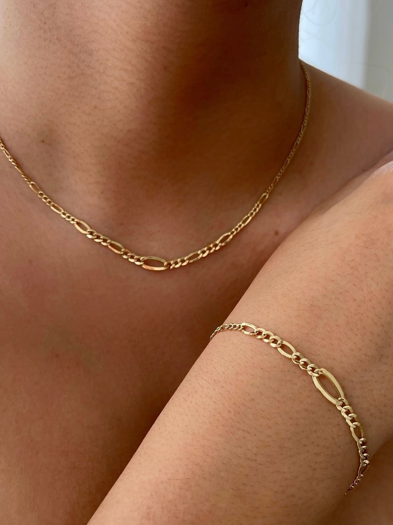 Nahaufnahme einer 18k Gelbgold Figaro-Kette und des passenden Goldarmbands, getragen auf Hals und Unterarm einer Person; feine, glänzende Oval- und Rundglieder, eleganter, minimalistischer Schmuckstil.
