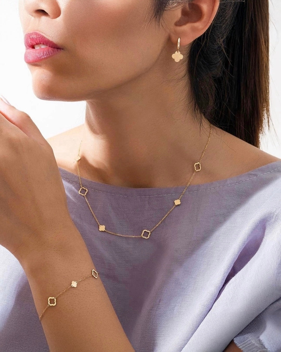 Frau trägt ein elegantes Schmuckset aus 18 Karat Gelbgold – bestehend aus Halskette, Armband und Ohrringen mit feinen Kleeblatt-Elementen. Der zarte Goldschmuck harmoniert mit ihrem fliederfarbenen Oberteil und unterstreicht einen femininen, modernen Look.