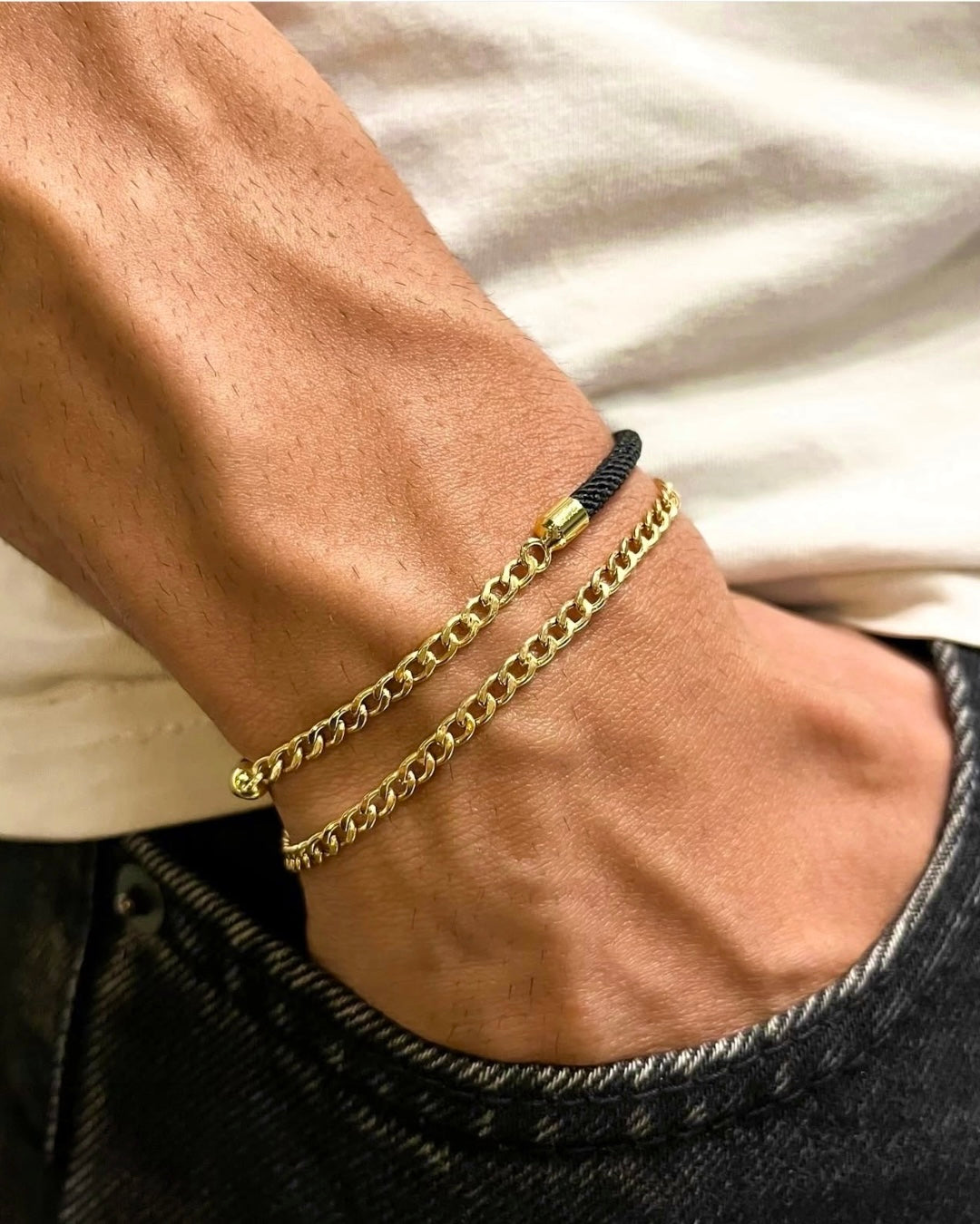 Goldarmband aus 18k Gelbgold im Cuban-Link-Stil am Handgelenk eines Mannes, kombiniert mit einem schwarzen Kordelarmband; Nahaufnahme mit warmem Licht, das die glänzenden, massiven Goldglieder und die elegante Struktur der Kette betont.