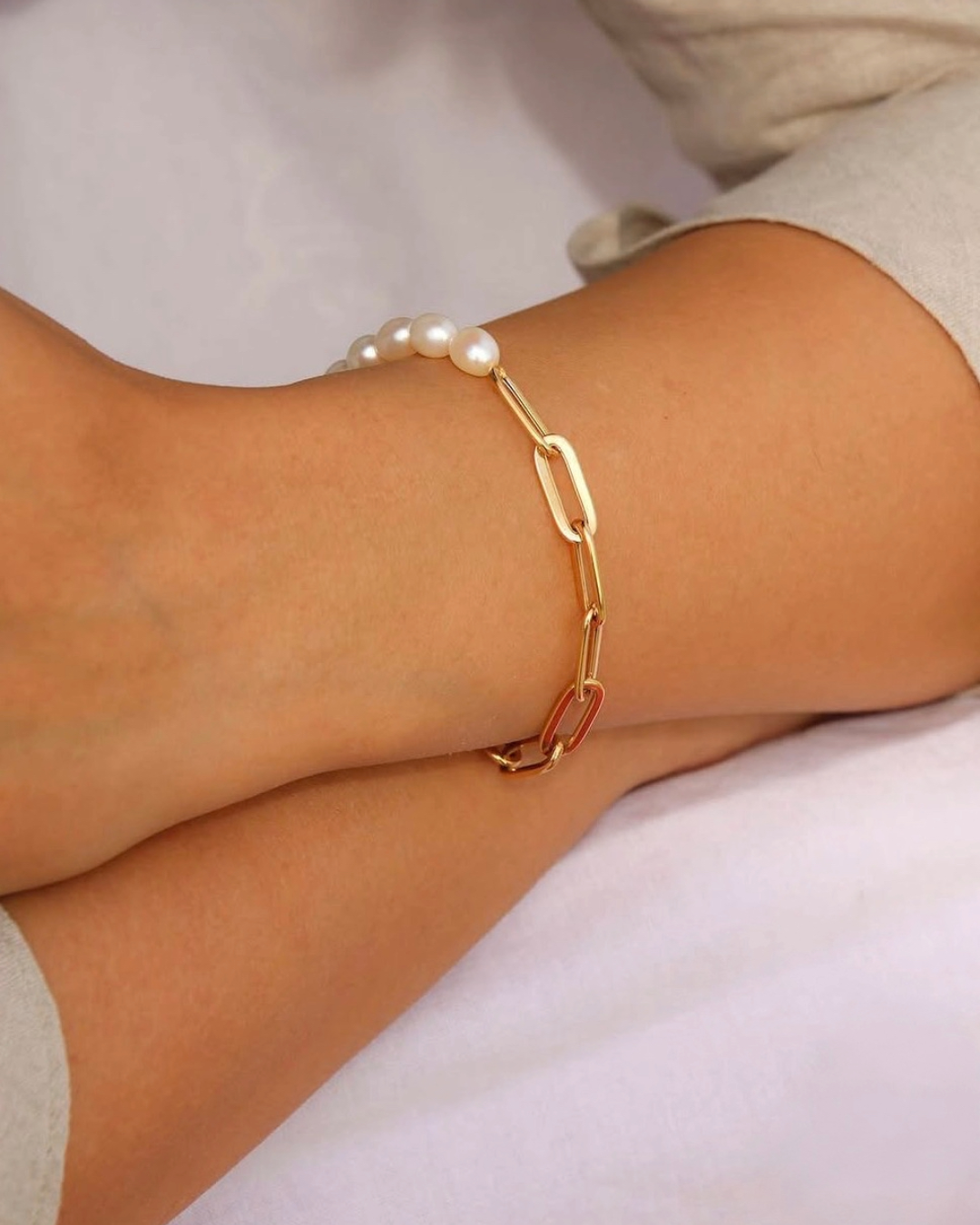 Nahaufnahme eines eleganten Armbands mit großen goldfarbenen Kettengliedern und weißen Perlen am Handgelenk. Das Schmuckstück kombiniert moderne Gliederoptik mit zeitloser Eleganz und wird in sanftem Licht stilvoll in Szene gesetzt. Minimalistische Luxusaufnahme mit hochwertiger, femininer Ausstrahlung.