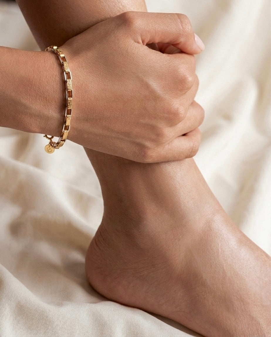 Nahaufnahme eines eleganten 18-karätigen Gelbgold-Armbands am Handgelenk einer Frau. Das Armband besteht aus rechteckigen Gliedern mit fein gravierten, geometrischen Mustern und polierten Goldflächen, die das Licht dezent reflektieren. Die Hand ruht entspannt auf dem Knöchel, während der helle, cremefarbene Stoffhintergrund eine warme und luxuriöse Atmosphäre schafft. Das Schmuckstück wirkt zeitlos, hochwertig und stilvoll.