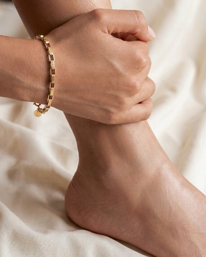 Nahaufnahme eines eleganten 18-karätigen Gelbgold-Armbands am Handgelenk einer Frau. Das Armband besteht aus rechteckigen Gliedern mit fein gravierten, geometrischen Mustern und polierten Goldflächen, die das Licht dezent reflektieren. Die Hand ruht entspannt auf dem Knöchel, während der helle, cremefarbene Stoffhintergrund eine warme und luxuriöse Atmosphäre schafft. Das Schmuckstück wirkt zeitlos, hochwertig und stilvoll.