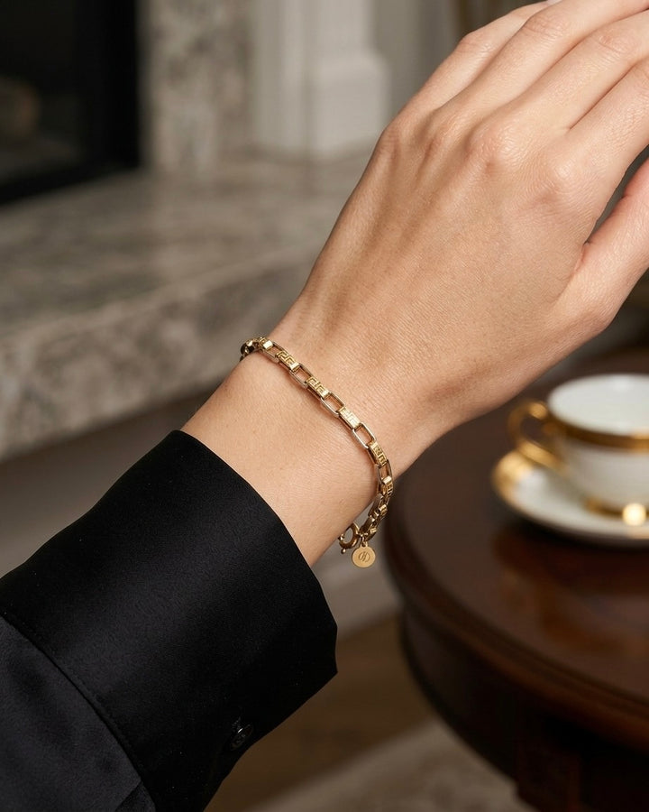 Elegante Lifestyle-Aufnahme eines filigranen 18-karätigen Bicolor-Armbands aus Gelb- und Weissgold am Handgelenk einer Frau. Die rechteckigen Glieder mit fein gravierten geometrischen Mustern verleihen dem Schmuckstück eine moderne und zugleich zeitlose Ausstrahlung. Die Trägerin trägt ein schwarzes, satinartiges Kleidungsstück, während im Hintergrund ein stilvoll gedeckter Tisch mit einer Tasse und Untertasse zu sehen ist. Die warme Atmosphäre unterstreicht den luxuriösen und anspruchsvollen Charakter des 