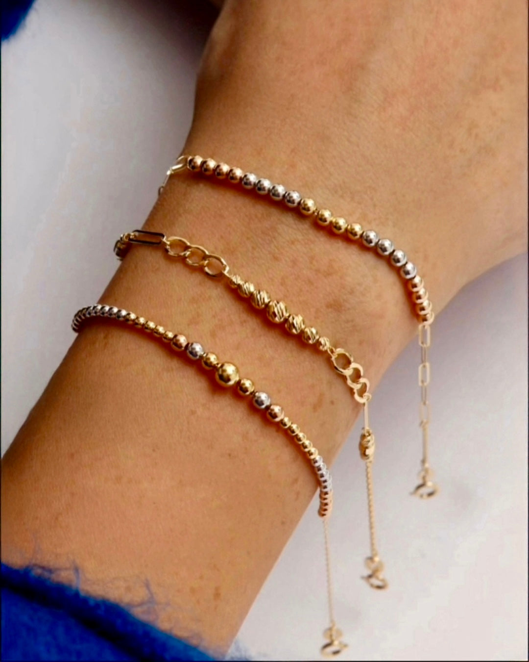 Drei feine Goldarmbänder aus 18k Gelbgold, Weissgold und Roségold am Handgelenk getragen – filigrane Kugelarmbänder mit polierter Oberfläche und zarten Verlängerungsketten, elegante Kombination in warmen Goldtönen.