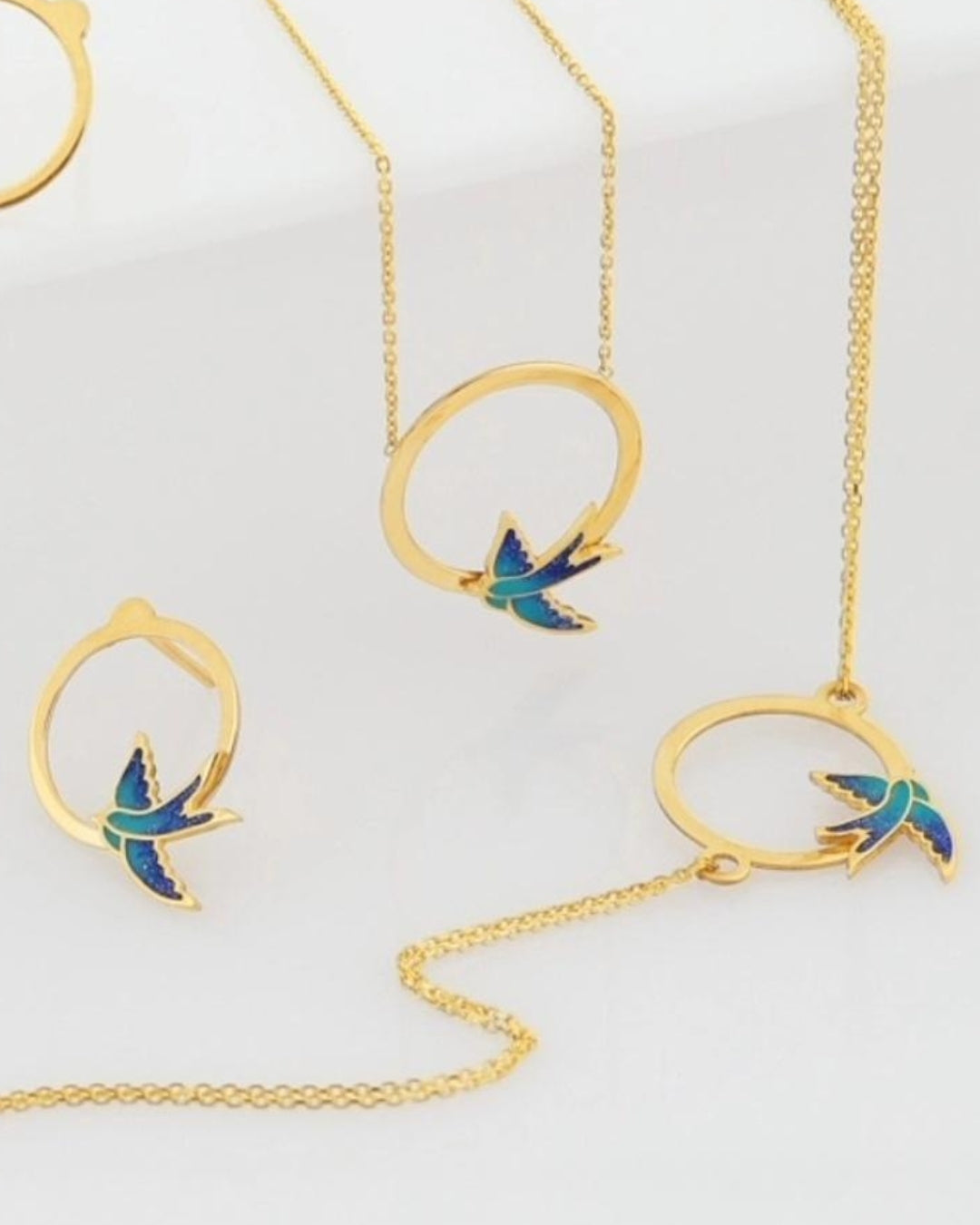 Nahaufnahme des 18k Gold Schmucksets Parastoo mit emaillierten Schwalben – zeigt feine Details von Kette, Armband &amp; Ohrsteckern.
