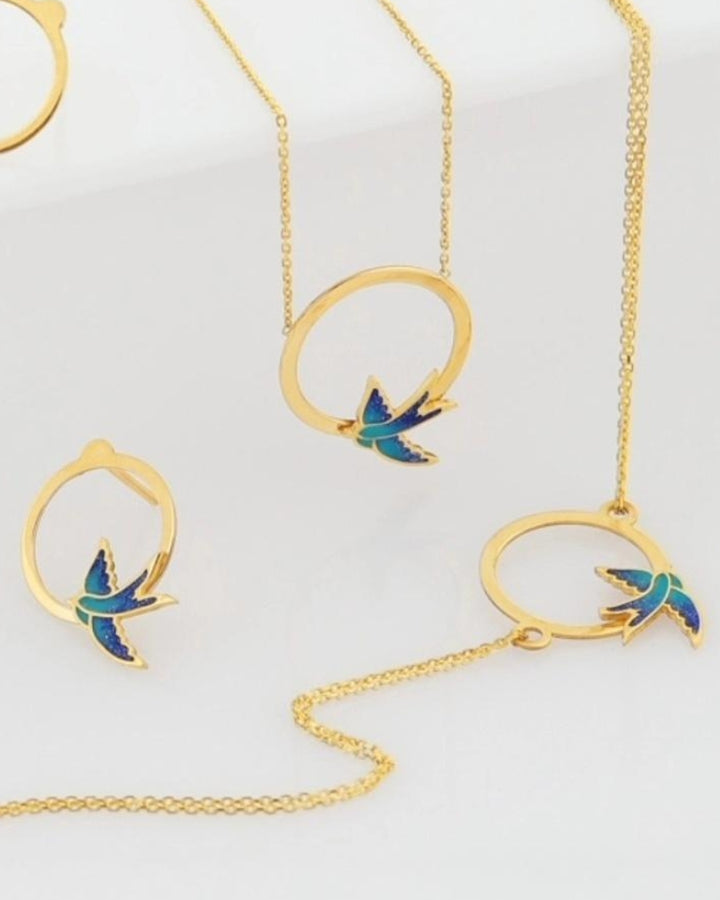 Nahaufnahme des 18k Gold Schmucksets Parastoo mit emaillierten Schwalben – zeigt feine Details von Kette, Armband &amp; Ohrsteckern.