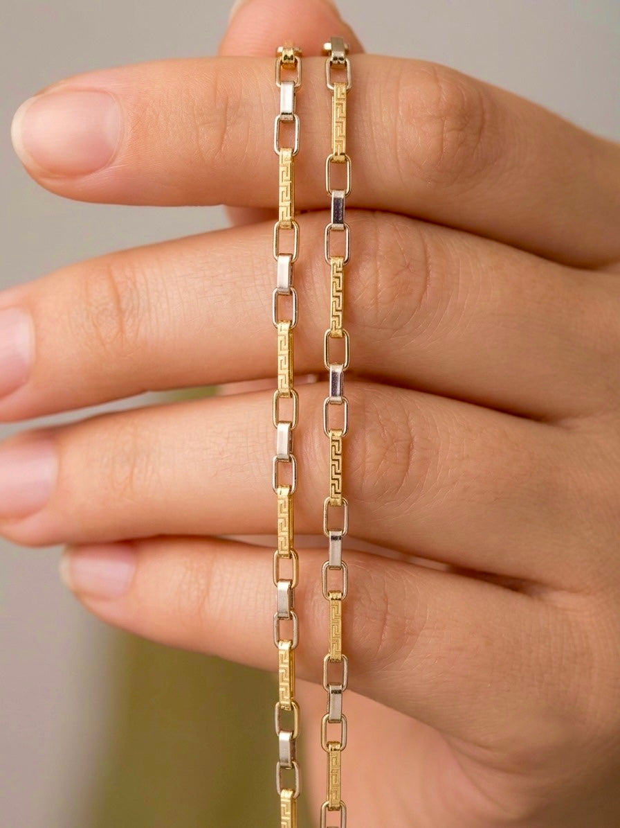 Nahaufnahme einer eleganten 18k Gelbgold-Halskette mit griechischem Mäander-Muster und weissgoldfarbenen Zwischengliedern. Die feingliedrige Kette wird auf einer Hand präsentiert und vereint zeitloses Design mit persisch inspirierter Handwerkskunst.