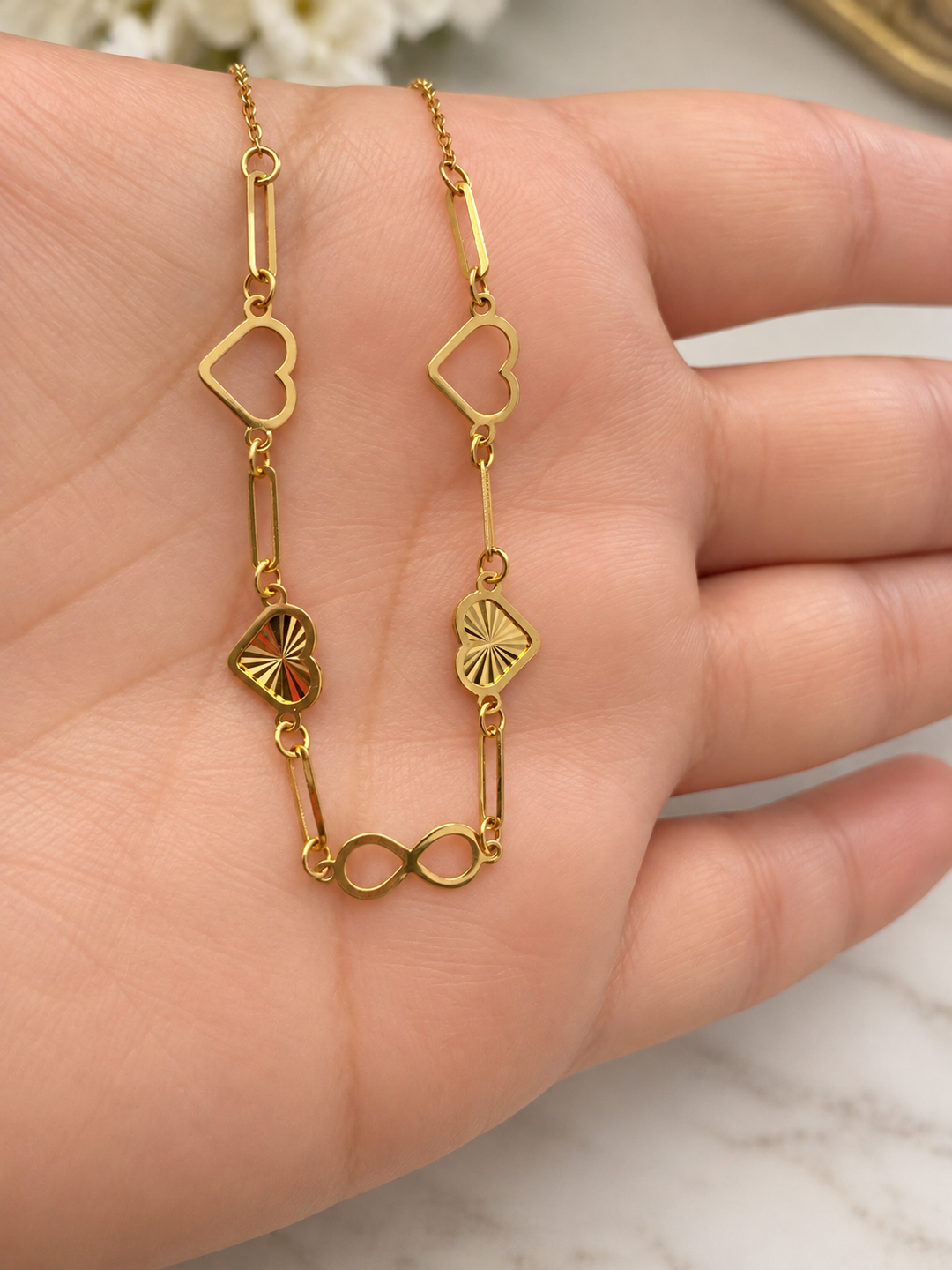 Nahaufnahme der goldenen Halskette Eshgh-e Bi Payan auf einer Hand mit offenen und strukturierten Herzen sowie Infinity-Symbol.