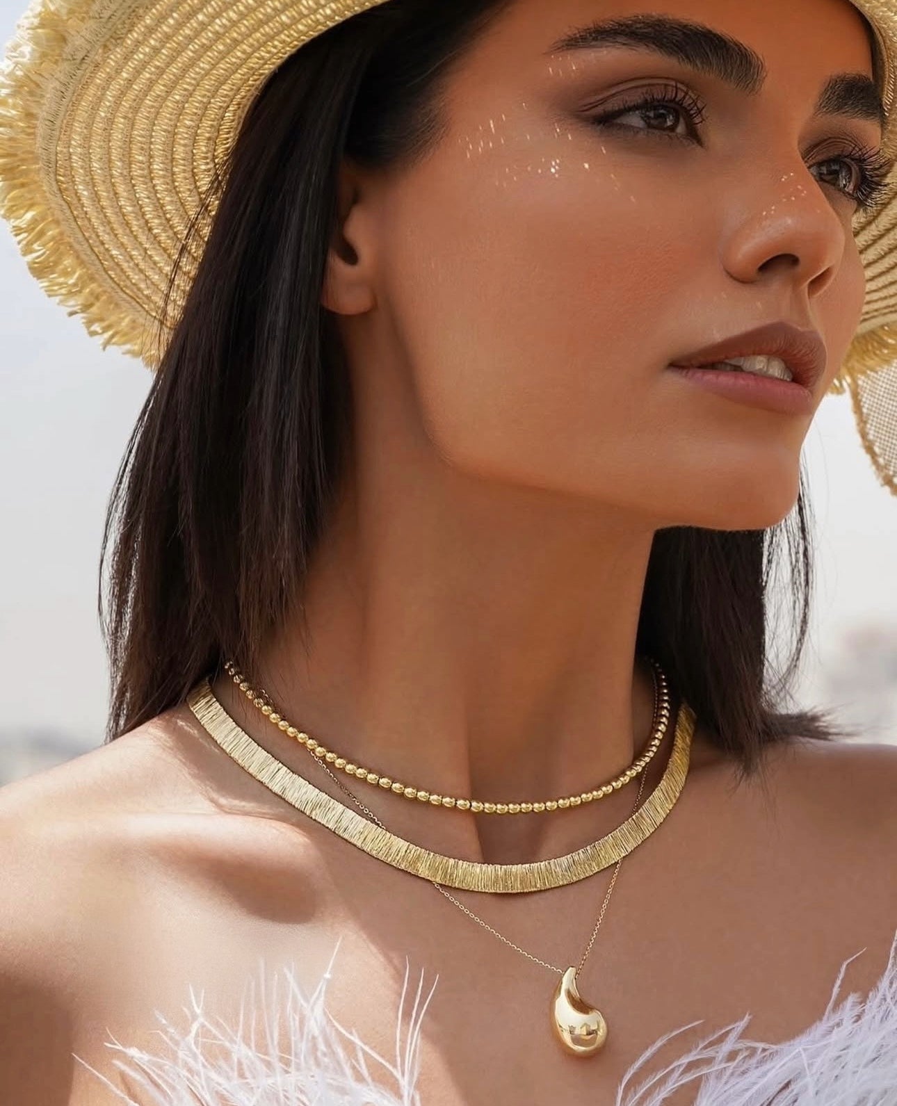 Nahaufnahme einer Frau mit Strohhut, die eine elegante 18k Gelbgold-Perlenkette dicht am Hals trägt, kombiniert mit einer breiten, strukturierten Goldcollier-Kette und einer feinen Kette mit tropfenförmigem Anhänger.