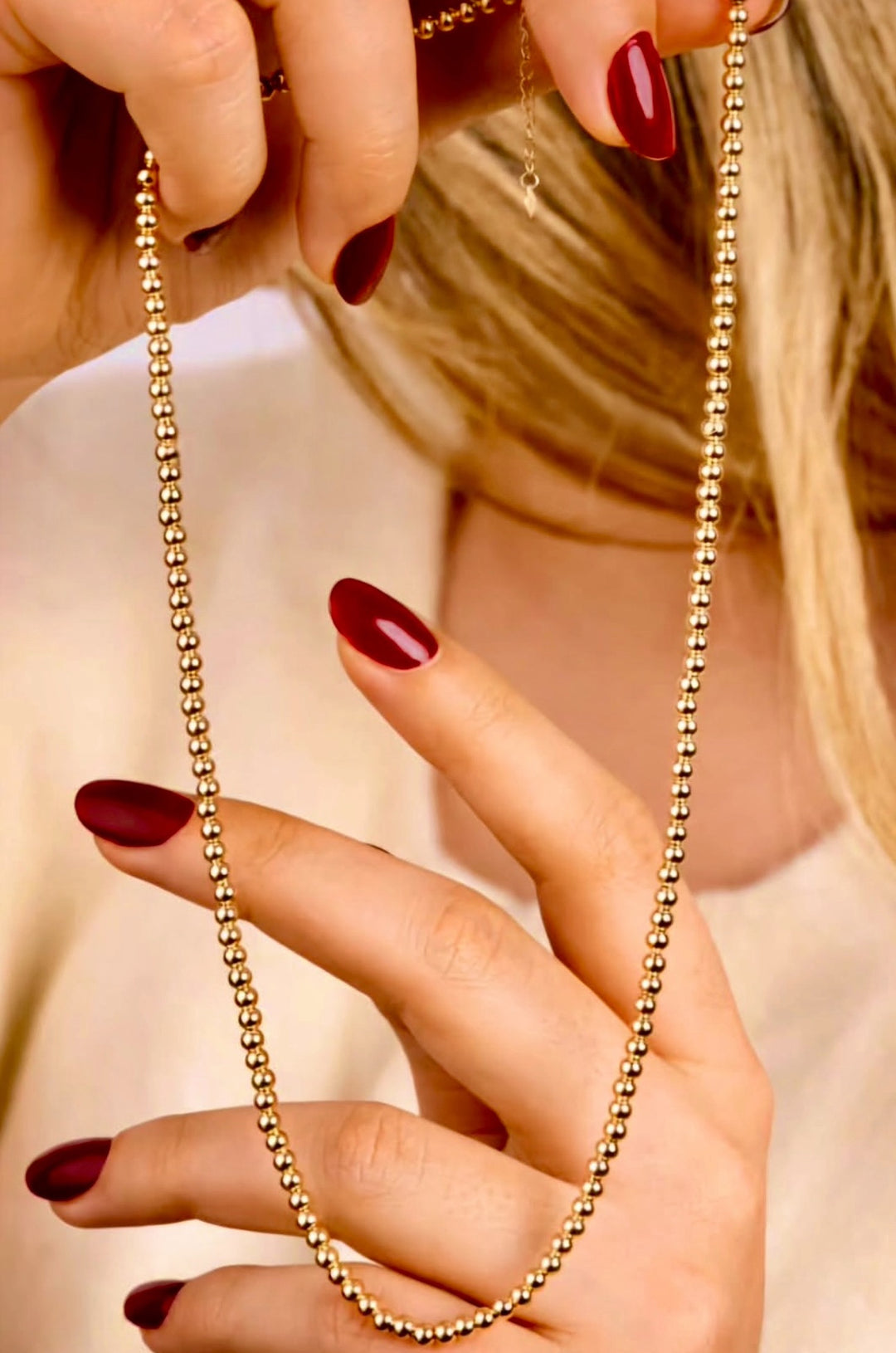 Nahaufnahme von Händen mit dunkelrotem Nagellack, die eine 18k Gelbgold-Perlenkette mit gleichmäßig angeordneten, polierten Goldkugeln halten, vor einem weichen, beigefarbenen Hintergrund.