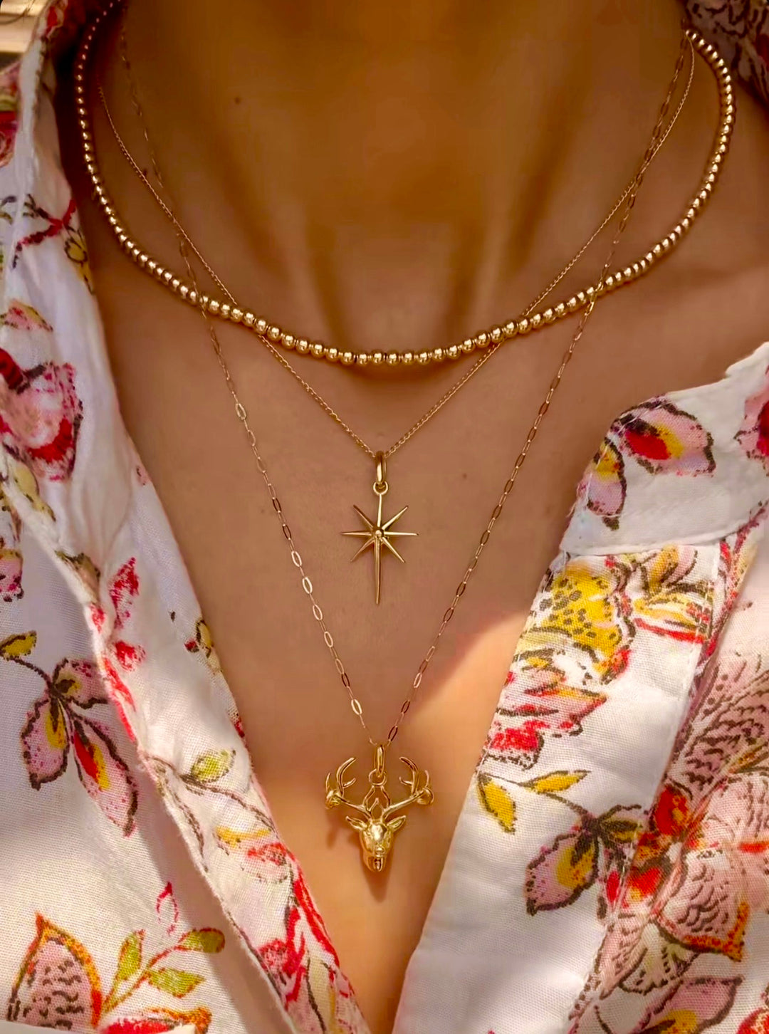 Nahaufnahme eines Halses mit drei übereinander getragenen Halsketten aus 18k Gelbgold. Die oberste Kette ist eine feine Perlenkette mit gleichmäßig runden, polierten Goldkugeln. Darunter hängt eine filigrane Kette mit einem achtzackigen Sternanhänger, und die längste Kette trägt einen detailreichen Hirschkopf-Anhänger. Getragen zu einer weißen Bluse mit farbigem Blumenmuster.