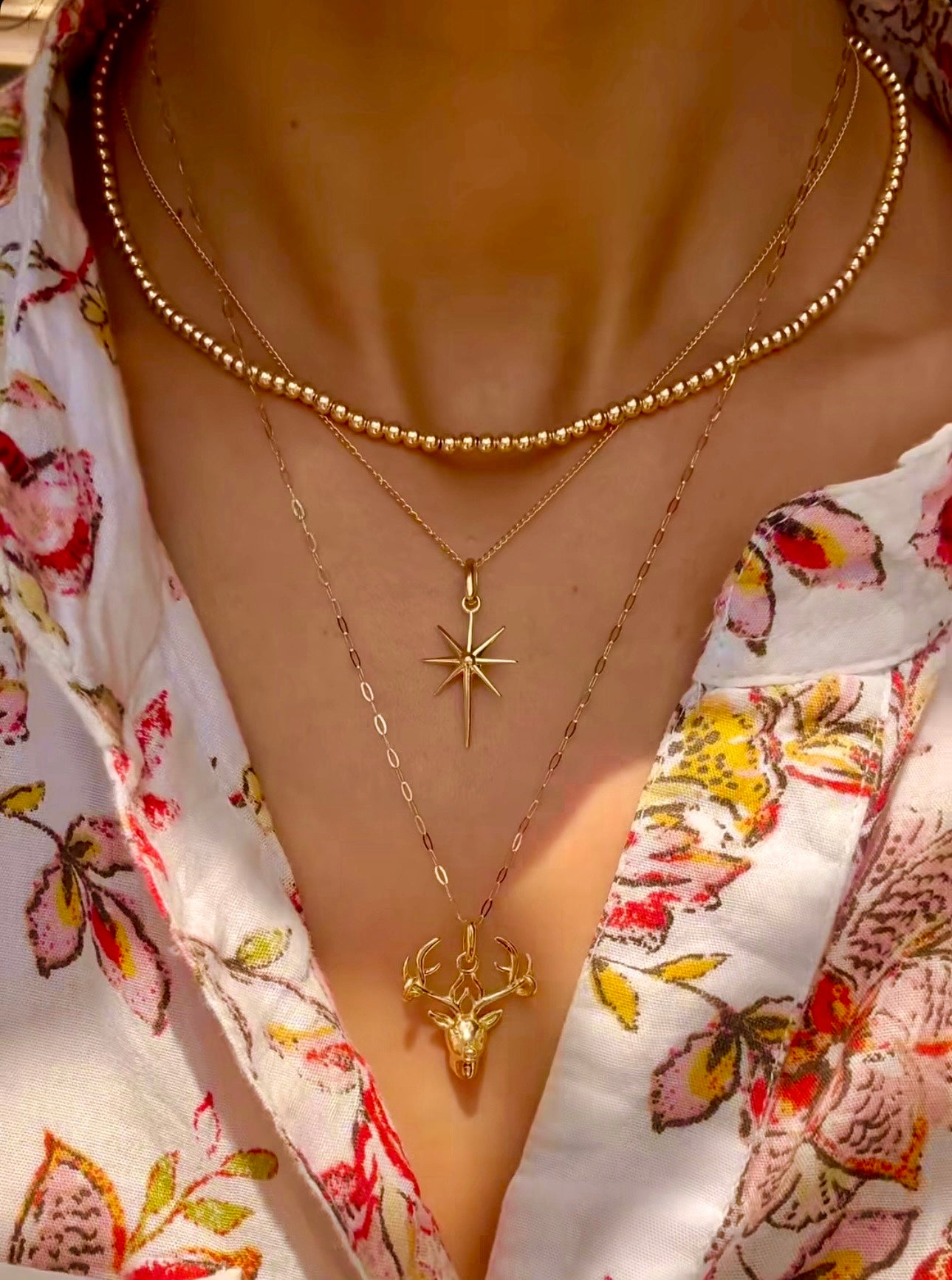 Nahaufnahme eines Halses mit drei übereinander getragenen Halsketten aus 18k Gelbgold. Die oberste Kette ist eine feine Perlenkette mit gleichmäßig runden, polierten Goldkugeln. Darunter hängt eine filigrane Kette mit einem achtzackigen Sternanhänger, und die längste Kette trägt einen detailreichen Hirschkopf-Anhänger. Getragen zu einer weißen Bluse mit farbigem Blumenmuster.