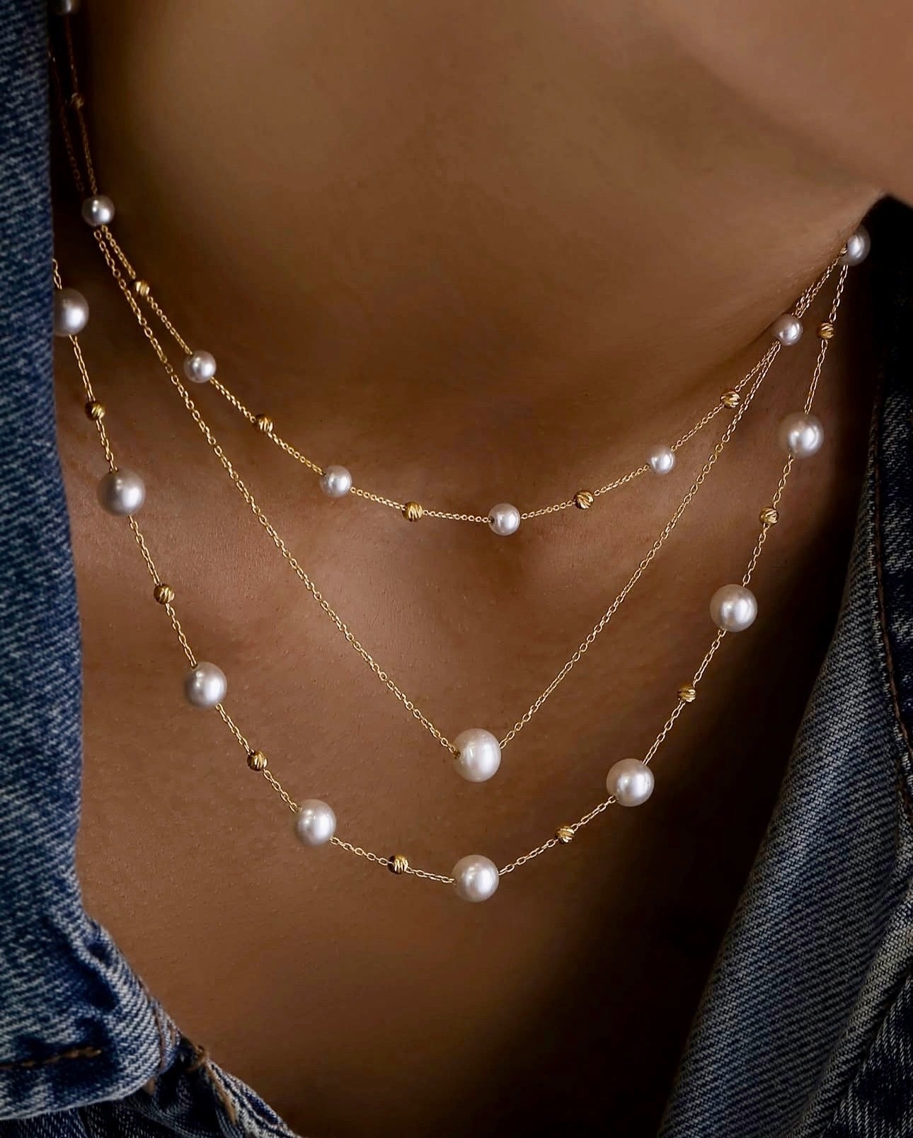Nahaufnahme eines Halses mit drei übereinander getragenen Halsketten aus 18k Gelbgold („Morvarid-e Noor“ – Lichterperlen). Die feinen Goldketten sind mit gleichmäßig verteilten weißen Perlen und kleinen, strukturierten Goldelementen verziert. Getragen zu einer blauen Jeansjacke im lässigen Stil.