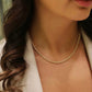 Nahaufnahme einer Frau mit lockigem, braunem Haar, die eine elegante Halskette aus 18k Gelbgold mit feiner, gedrehter Gliederstruktur trägt. Sie trägt ein helles Oberteil und ein cremefarbenes Blazer-Jackett, aufgenommen bei weichem, natürlichem Licht.