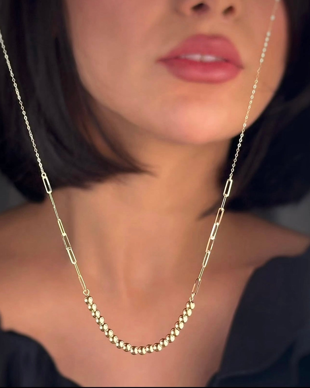 Nahaufnahme einer Frau, die eine elegante 18k-Gelbgold-Halskette trägt. Die Kette kombiniert fein gearbeitete, hochglanzpolierte Goldkugeln im Mittelteil mit länglichen, modernen Kettengliedern. Das Schmuckstück liegt sanft am Dekolleté an und wirkt minimalistisch, hochwertig und zeitlos vor dunklem Hintergrund.
