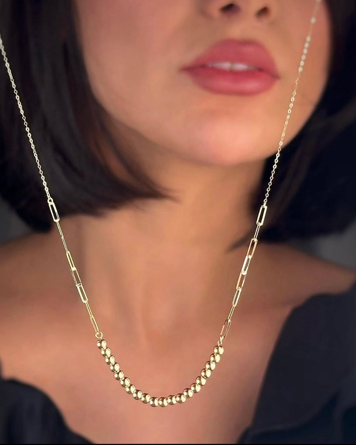 Nahaufnahme einer Frau, die eine elegante 18k-Gelbgold-Halskette trägt. Die Kette kombiniert fein gearbeitete, hochglanzpolierte Goldkugeln im Mittelteil mit länglichen, modernen Kettengliedern. Das Schmuckstück liegt sanft am Dekolleté an und wirkt minimalistisch, hochwertig und zeitlos vor dunklem Hintergrund.