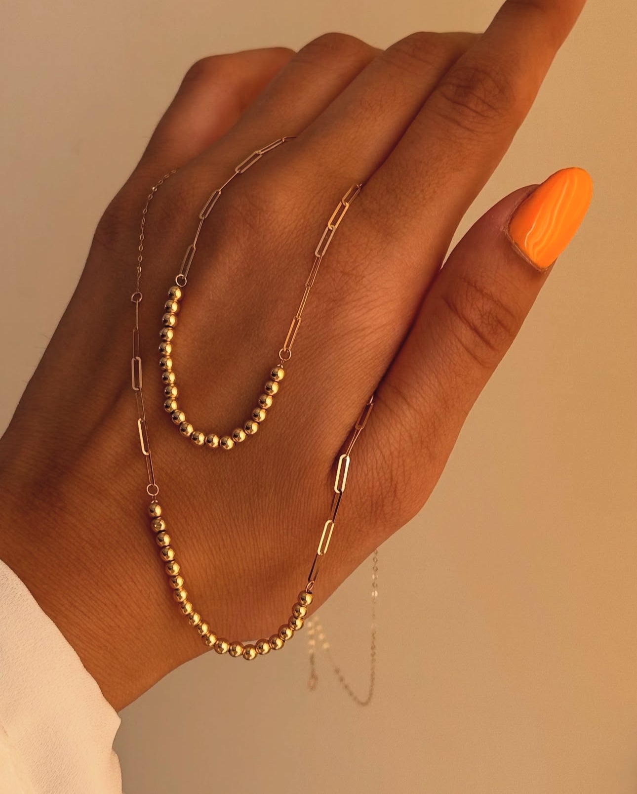 Nahaufnahme einer Goldperlenkette aus 18k Gelbgold, locker über eine Hand mit orange lackiertem Daumennagel gelegt. Die Kette kombiniert polierte Goldperlenabschnitte mit länglichen, feingliedrigen Kettengliedern, was ihr einen modernen, verspielten Look verleiht. Hintergrund in warmem Beigeton.