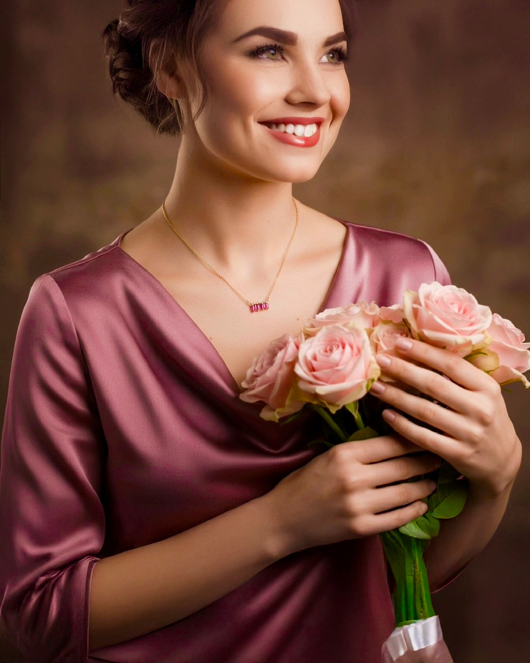 Frau in elegantem Seidenkleid mit rosafarbenem Rosenstrauss, trägt eine filigrane 18k Gelbgold-Halskette mit rosa Edelsteinen – zeitloser Goldschmuck von Ziba Gold