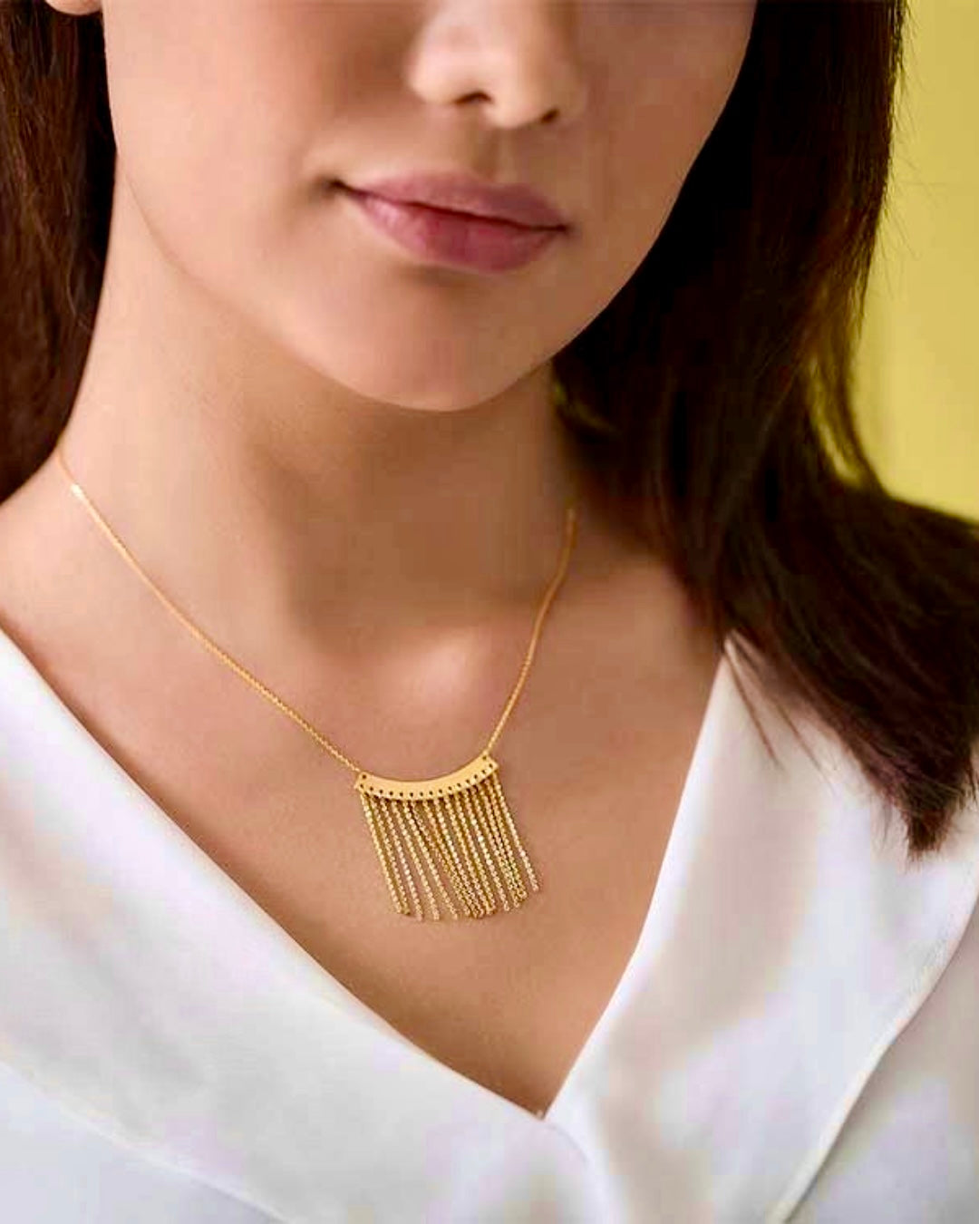 Nahaufnahme einer Frau mit einer Halskette aus 18k Gelbgold. Der Anhänger „Baran-e Zarrin“ (Goldener Regen) besteht aus einer gebogenen Goldplatte, von der mehrere feine Goldketten wie Regentropfen herabhängen. Getragen zu einer eleganten weißen Bluse vor gelbem Hintergrund.
