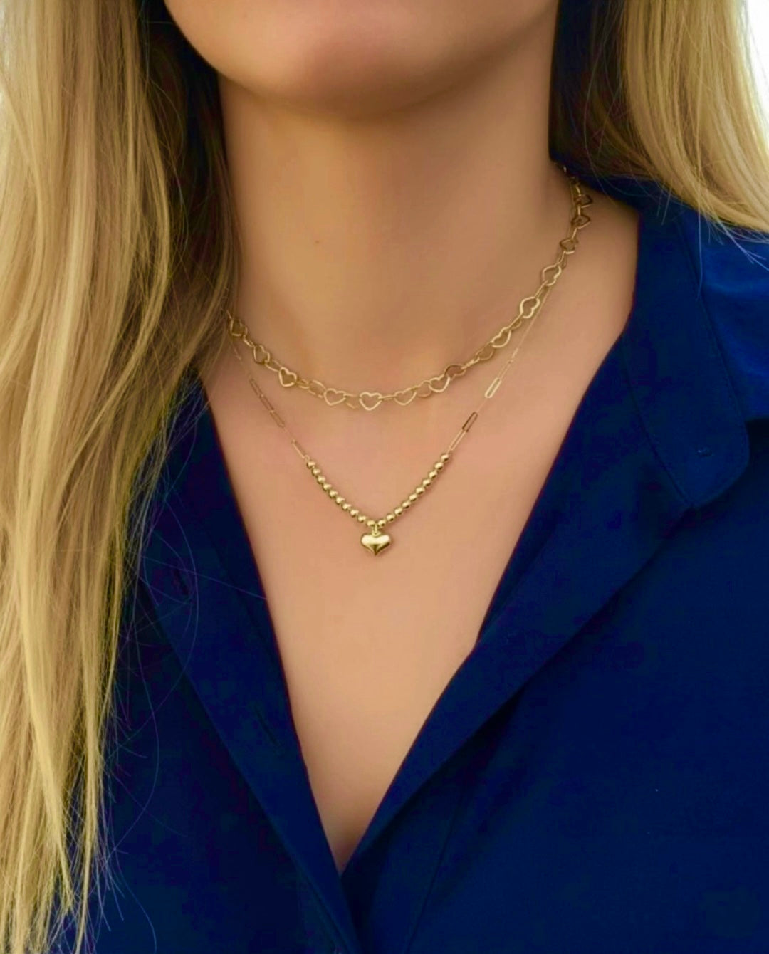 Modell trägt zwei filigrane 18k-Gelbgold-Halsketten: oben eine feine Kette mit kleinen Herzgliedern, darunter eine zarte Kugelkette mit glänzendem Herzanhänger, elegant kombiniert zu einem Layering-Look.