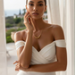 Elegantes Portrait einer stilvollen Frau in einem schulterfreien weißen Kleid vor hellem, mediterranem Hintergrund. Sie trägt eine filigrane Goldhalskette mit mehreren polierten Goldkugeln als zentrales Element. Das warme Licht betont ihre glatte Haut, die feinen Gesichtszüge und die luxuriöse, minimalistische Ästhetik des Schmucks. Die Szene wirkt hochwertig, ruhig und exklusiv.